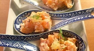 Four decorative ceramic spoons with diced raw salmon, onions, and herbs, garnished with dill and pepper, arranged neatly on a rectangular plate.