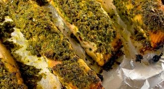 Herb-crusted salmon fillets baked on a sheet of aluminium foil, with a golden green topping and slightly crispy edges. The fish appears moist and well-seasoned.