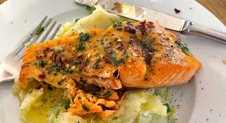 A plate of mashed potatoes topped with a seasoned, baked salmon fillet, garnished with herbs and spices. A fork and knife rest on the plate, and a glass is visible in the background on a wooden table.