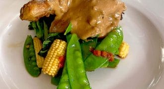 A roasted chicken leg topped with creamy brown sauce, served on a bed of sautéed mangetout, baby sweetcorn, and greens on a white plate.
