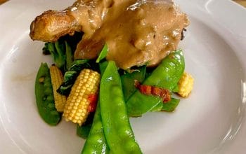 A roasted chicken leg topped with creamy brown sauce, served on a bed of sautéed mangetout, baby sweetcorn, and greens on a white plate.