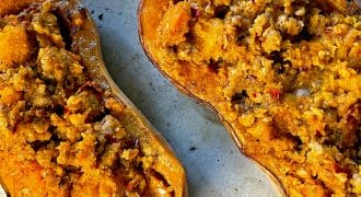 Two halves of butternut squash are filled with a crumbly, golden brown stuffing and baked on a metal tray. The stuffing appears to include grains and vegetables, giving a textured appearance.