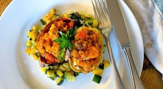 A white plate with stuffed peppers filled with a vegetable and potato mixture, garnished with parsley, and surrounded by diced sautéed courgette. A fork and knife rest at the side of the plate, on a wooden table.