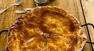 A golden-brown, freshly baked pie in a tin sits on a rustic wooden table, with two spoons and jars of condiments in the background. The pie has a flaky, glossy pastry lid and a single cut starting from the centre.