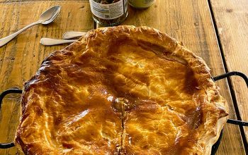 A golden-brown, freshly baked pie in a tin sits on a rustic wooden table, with two spoons and jars of condiments in the background. The pie has a flaky, glossy pastry lid and a single cut starting from the centre.