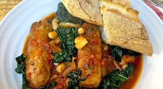 A white plate with sausage stew, featuring sausages, leafy greens, beans, and tomato sauce, garnished with two pieces of crusty bread on top. A fork and spoon rest on a serviette nearby.