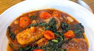 A white bowl filled with sausage stew, featuring sausages, cherry tomatoes, greens, and beans in a tomato-based sauce. A crusty piece of bread sits on a small black plate in the background.