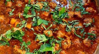 A close-up of a pot filled with a chunky tomato-based stew, featuring pieces of meat, vegetables, and topped with fresh chopped herbs.