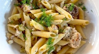 A white bowl filled with penne pasta, meatballs, creamy sauce, and garnished with fresh herbs, sitting on a wooden table.