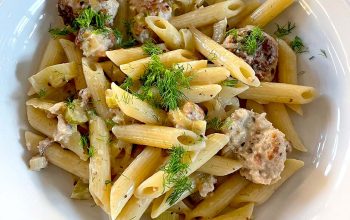 A white bowl filled with penne pasta, meatballs, creamy sauce, and garnished with fresh herbs, sitting on a wooden table.