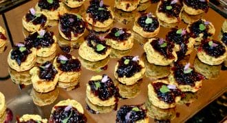 A tray filled with small, round appetiser bites topped with a dark jam or chutney and garnished with tiny herbs and edible flowers, arranged neatly for serving.