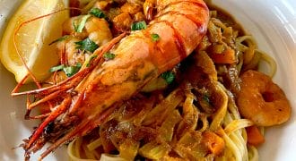 A bowl of pasta topped with a large grilled king prawn, creamy tomato sauce, chopped herbs, and a lemon wedge on the side. Prawn pieces and diced vegetables are mixed in with the pasta.