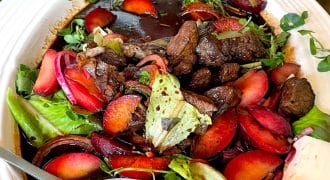 A white bowl filled with a colourful salad of leafy greens, sliced plums, seared beef pieces, and a dark balsamic dressing, with a large serving spoon resting inside the bowl.