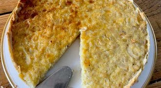 A partially sliced onion tart sits on a white plate atop a wooden table, with a metal serving spatula resting nearby. The tart has a golden-brown, slightly uneven surface and a flaky pastry.