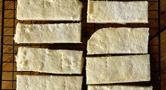 Rectangular pieces of golden, sugar-dusted shortbread are cooling on a black wire rack set over a wooden surface. The shortbread bars are arranged neatly in rows.