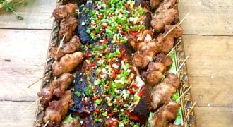 A large tray of grilled meat skewers arranged along the sides, with assorted grilled meats in the centre topped with chopped spring onions and red chillies, displayed on a wooden table.