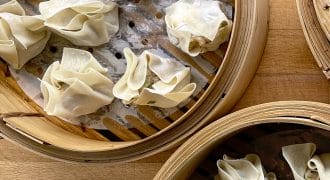 Three bamboo steamers filled with uncooked dumplings, arranged on a wooden surface. The dumplings are wrapped in thin pastry and placed on greaseproof paper inside the steamers.