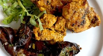 Grilled pieces of seasoned chicken served with roasted aubergine garnished with herbs and a small side of fresh rocket on a white plate.