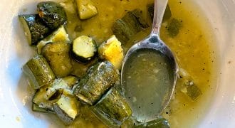 A white bowl containing chunks of cooked courgette in a yellow-green broth, with a metal spoon resting in the bowl. Some broth and vegetable pieces are scattered around.
