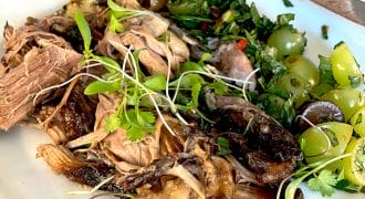 A plate of shredded cooked meat with microgreens, roasted vegetables, sliced green grapes, and fresh herbs, served on a white dish on a wooden table. A knife rests on the plate’s edge.