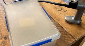 A plastic container with a blue catch sits on a wooden table beside a silver handheld kitchen tool with a black hose attached. A piece of crumpled paper is also on the table near the container.