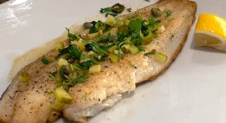 A cooked fish fillet garnished with chopped herbs and capers is served on a white plate with a lemon wedge on the side. A hand holds the plate, and part of a patterned garment is visible in the background.