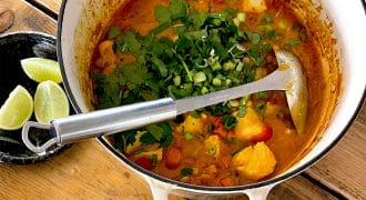 A pot of fish stew with chunks of fish, vegetables, and fresh herbs sits on a wooden table next to a bowl of white rice and a plate with lime wedges. A ladle rests inside the pot.