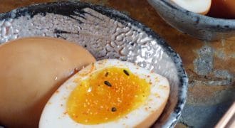 A ceramic bowl with a halved soft-boiled egg, seasoned with spices and black sesame seeds. Another bowl contains whole and halved eggs. Chopsticks are placed nearby on a stone surface.