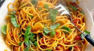 A bowl of spaghetti with tomato sauce, fresh basil leaves, and a pasta serving spoon, sitting on a wooden surface. Steam rises from the pasta, indicating it is freshly cooked.