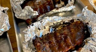 Barbecued ribs covered in sauce, partially wrapped in aluminium foil, served on metal trays on a wooden table. The ribs appear juicy and well-cooked, ready to be eaten.