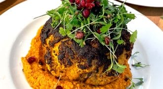 A whole roasted cauliflower sits on a bed of orange sauce, garnished with fresh microgreens and pomegranate seeds, served on a white plate.