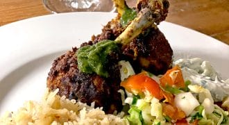 A plate of spiced chicken drumsticks topped with green sauce, served with white rice, chopped vegetable salad, and a side of creamy sauce, with a wine glass in the background.