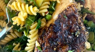 Close-up of a plate with seasoned grilled chicken breast, rotini pasta, leafy greens, peas, and vegetables, all mixed together. A fork is partially visible on the left side of the dish.