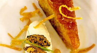 A slice of golden syrup cake garnished with thin orange zest strips, served with a scoop of vanilla ice cream and caramelised orange slices on a white plate.