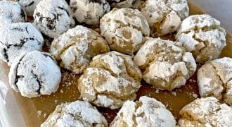 A container lined with brown baking parchment holds rows of crinkle biscuits. The biscuits are covered in icing sugar; some are chocolate and others are light-coloured, suggesting different flavours.