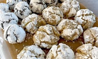 A container lined with brown baking parchment holds rows of crinkle biscuits. The biscuits are covered in icing sugar; some are chocolate and others are light-coloured, suggesting different flavours.