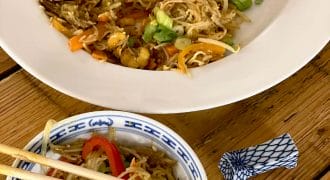 A large white plate and a small patterned bowl filled with stir-fried noodles, vegetables, and spring onions sit on a wooden table. Chopsticks rest on the bowl, with a chopstick holder nearby.