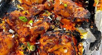 Charred spicy baked chicken wings on a foil-lined tray, garnished with sliced spring onions and sesame seeds. The wings are coated in a red sauce, with crispy edges and visible bits of seasoning.