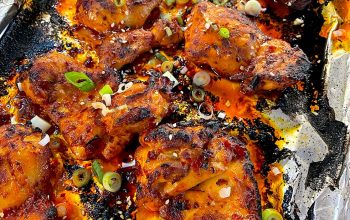 Charred spicy baked chicken wings on a foil-lined tray, garnished with sliced spring onions and sesame seeds. The wings are coated in a red sauce, with crispy edges and visible bits of seasoning.