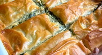 A close-up of golden-brown, flaky spanakopita cut into rectangles in a white baking dish, with crispy filo pastry visible on top and edges.