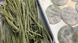 Fresh green pasta dough cut into fettuccine strips sits in one tray, while round green ravioli wrappers dusted with flour are arranged on another tray. Both trays are on a wooden surface.
