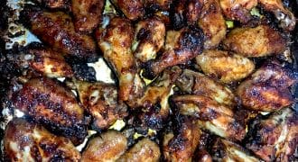 A close-up view of a baking tray filled with numerous baked chicken wings, golden-brown and caramelised, with crispy, charred edges.