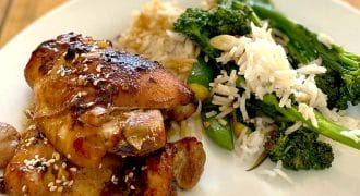 A plate of glazed chicken thighs sprinkled with sesame seeds, served with white rice and sautéed green vegetables, including tenderstem broccoli, on a white plate on a wooden table.