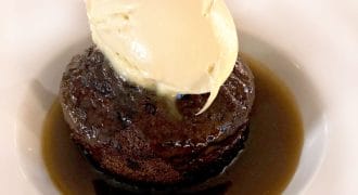 A dessert of sticky toffee pudding sits in a pool of toffee sauce on a white plate, topped with a scoop of vanilla ice cream.