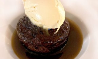 A dessert of sticky toffee pudding sits in a pool of toffee sauce on a white plate, topped with a scoop of vanilla ice cream.