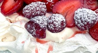 A close-up of a pavlova dessert topped with whipped cream, fresh strawberries, raspberries, and a dusting of icing sugar on a white plate.