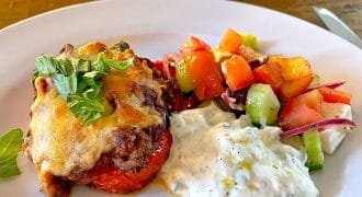 A white plate with a serving of baked dish topped with melted cheese and herbs, a scoop of creamy tzatziki, and a colourful Greek salad with tomatoes, cucumber, red onion, and feta cheese. A baking dish is visible in the background.