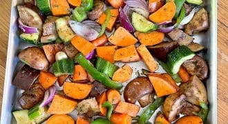A baking tray filled with chopped mixed vegetables including sweet potatoes, courgette, aubergine, carrots, green pepper, red onion, mushrooms, and whole garlic cloves, ready for roasting.