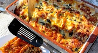 A baked lasagne in a rectangular dish with a knife inserted, surrounded by golden melted cheese and herbs. A plate with slices of crusty bread and a black spatula sits nearby on a sunlit wooden table.