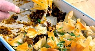 A person serves a portion of a baked dish with golden, crispy filo pastry on top, revealing a rich, dark filling underneath in a white rectangular baking dish. Parsley is sprinkled on the pastry.